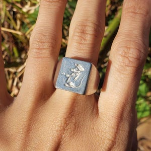 May include: A silver signet ring with a square face featuring an engraved image of a mouse wearing a hat.