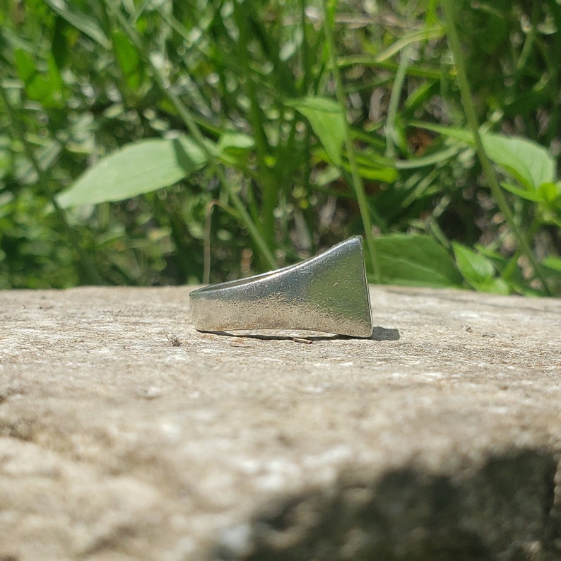 May include: A silver ring with a flat, triangular top. The ring is sitting on a rough, gray surface.