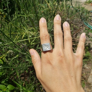 May include: A silver signet ring with a raised design on the top. The ring is worn on the index finger of a person's hand.