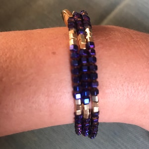 Può includere: Un braccialetto a treccia con perline dorate e viola. Le perline sono quadrate e hanno una finitura metallica.