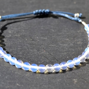 Può includere: Un braccialetto con perline di opale bianco, un cordoncino blu e una chiusura a scorrimento argentata. Il braccialetto è regolabile e può essere indossato al polso o alla caviglia.