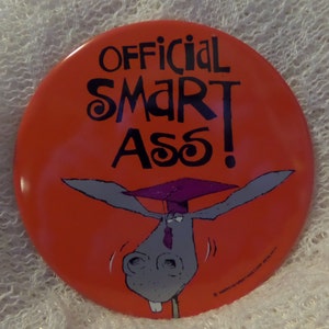 May include: A round, orange button with the words "OFFICIAL SMART ASS!" in black, stylized lettering. A cartoon donkey wearing a graduation cap is featured below the text. The button is a humorous novelty item.