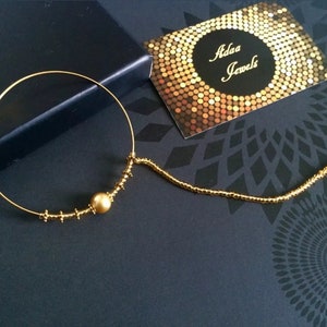 Op de afbeelding: Gouden neusring met een kleine parel en een eraan bevestigde ketting. De ring ligt op een zwarte achtergrond met een visitekaartje waarop "Adaa Jewels" staat.