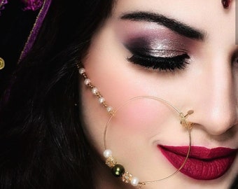 May include: A gold nose ring with a pearl and green bead chain. The nose ring is worn by a person with dark hair and makeup. The person is wearing a red lipstick.