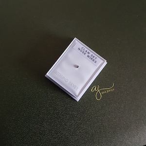 May include: A small, claw-set nose wire, displayed on a white card within a white box. The card reads "CLAW SET NOSE WIRES" and "STERLING SILVER". The background is a dark green surface, with a gold signature logo in the corner.