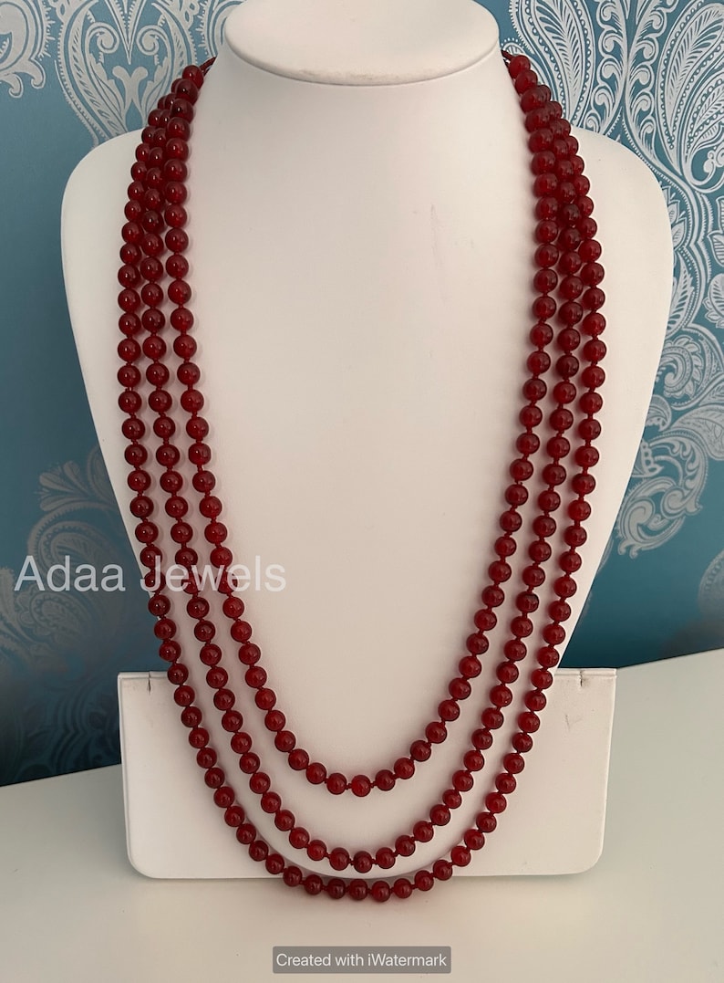 K&ouml;nnte beinhalten: Eine dreireihige Halskette aus dunkelroten Perlen. Die Halskette wird auf einer wei&szlig;en Mannequin-B&uuml;ste pr&auml;sentiert. Der Text "Adaa Jewels" ist in der linken unteren Ecke des Bildes sichtbar.
