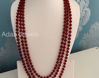 May include: A long, three-strand necklace made of dark red beads. The necklace is displayed on a white mannequin bust.