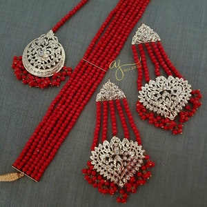 May include: A red beaded necklace set with gold and silver accents. The set includes a choker necklace, a pair of earrings, and a maang tikka.