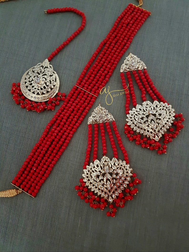 May include: A red beaded necklace set with gold and silver accents. The set includes a choker necklace, a pair of earrings, and a maang tikka.