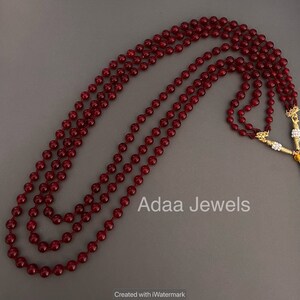 K&ouml;nnte beinhalten: Eine dreireihige Halskette aus dunkelroten Perlen. Die Halskette liegt auf einem grauen Hintergrund. Der Text "Adaa Jewels" ist auf dem Bild zu sehen.