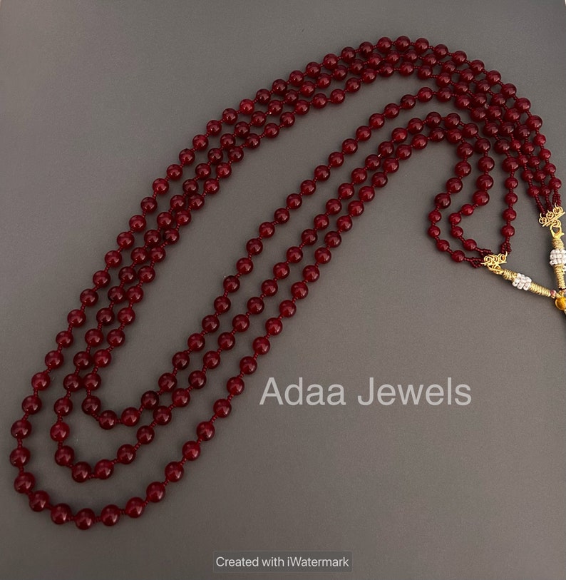 K&ouml;nnte beinhalten: Eine dreireihige Halskette aus dunkelroten Perlen. Die Halskette liegt auf einem grauen Hintergrund. Der Text "Adaa Jewels" ist auf dem Bild zu sehen.