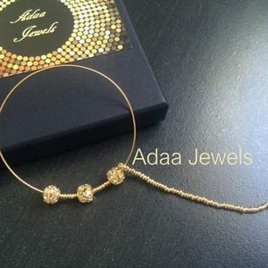 Op de afbeelding: Een gouden neusring met een eraan bevestigde ketting. De ring heeft drie ronde, heldere kristallen. De ketting is gemaakt van kleine gouden kralen. De neusring wordt getoond op een zwart oppervlak met een doos waarop "Adaa Jewels" staat.