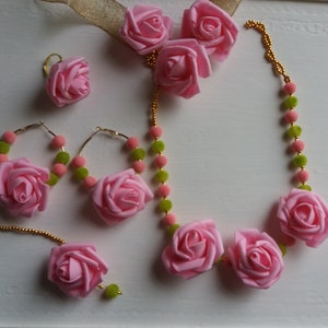May include: A pink flower jewelry set with a necklace, earrings, and a nose ring. The necklace is made of pink flowers and green beads. The earrings are hoop earrings with pink flowers and green beads. The nose ring is a single pink flower.