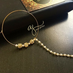 Op de afbeelding: Gouden neusring met een ketting van witte parels en drie ronde, glinsterende gouden kralen. De neusring ligt op een zwarte achtergrond met een visitekaartje waarop "aj adaa jewels" staat.