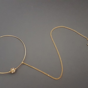 Op de afbeelding: Een gouden neusring met een klein, rond, helder kristal in het midden. De ring is bevestigd aan een gouden ketting die eindigt in een kleine, gouden haak.