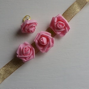 May include: Four pink fabric roses with gold trim, arranged on a gold glitter ribbon.