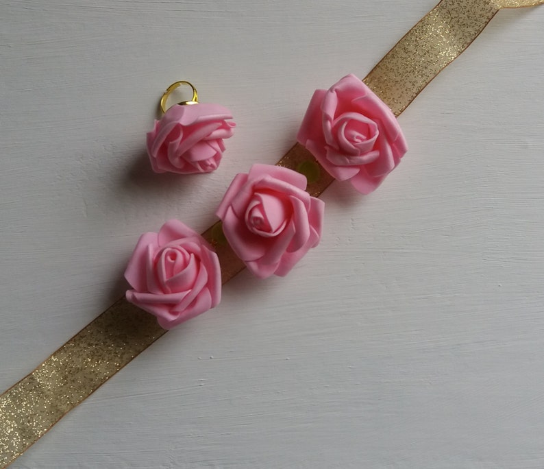 May include: Four pink fabric roses with gold trim, arranged on a gold glitter ribbon.