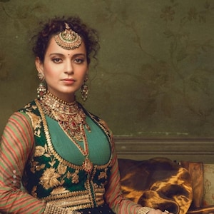 May include: A woman wearing a green and gold embroidered waistcoat over a teal top, with a gold and red necklace and matching earrings. She has a gold and green headpiece with a large jewel in the centre.