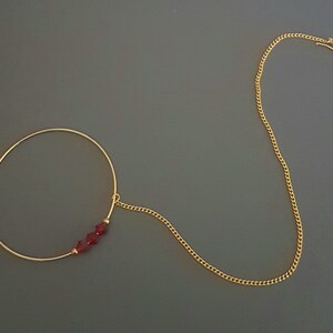 May include: A gold nose ring with a chain attached. The ring has three red beads on it.
