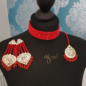 May include: A red beaded choker necklace with a matching pair of long earrings and a pendant. The earrings and pendant feature gold-toned metal accents with clear stones. The text "aj adam jewels" is visible in the image.