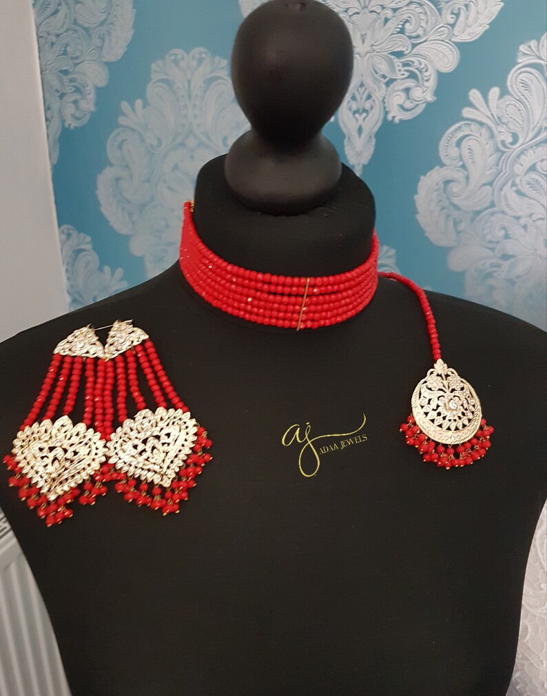 May include: A red beaded choker necklace with a matching pair of long earrings and a pendant. The earrings and pendant feature gold-toned metal accents with clear stones. The text "aj adam jewels" is visible in the image.
