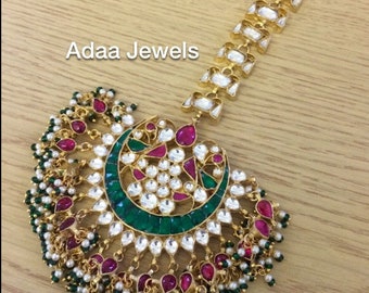May include: An ornate gold-toned headpiece with a crescent-shaped design, featuring green, red, and clear gemstones. The headpiece is adorned with small white and green beads. The text "Adaa Jewels" is visible.