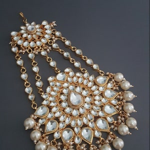 May include: A gold and white bridal hair accessory with a large, ornate, circular design featuring clear stones and pearls. The accessory has a chain with multiple strands and a smaller, circular design at the top.