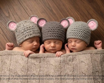May include: Three infants are wearing gray crocheted hats with round ears featuring pink centers. The hats have a textured knit pattern. The infants are lying side by side and are covered by a large, beige, cable-knit blanket with a visible texture.