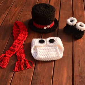 May include: A crocheted snowman costume set for a baby. The set includes a white crocheted nappy cover with black buttons, a black crocheted top hat with a red and white trim, a red crocheted scarf, and a pair of white and black crocheted booties.
