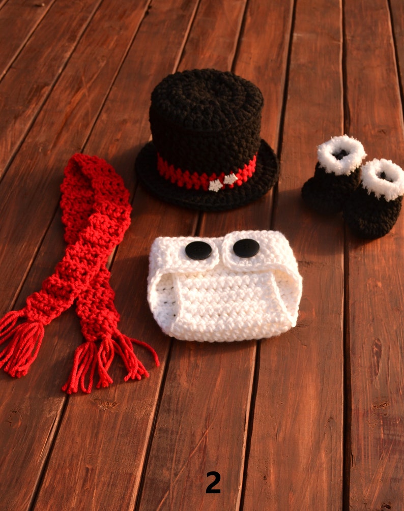May include: A crocheted snowman costume set for a baby. The set includes a white crocheted nappy cover with black buttons, a black crocheted top hat with a red and white trim, a red crocheted scarf, and a pair of white and black crocheted booties.