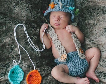 May include: A newborn baby wearing a blue crocheted fishing hat with colorful embellishments, a crocheted blue and gray fishing outfit, and holding a white string with two crocheted fish, one blue and one orange.