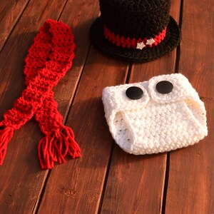 May include: A white crocheted nappy with two black buttons, a red crocheted scarf with tassels, and a black crocheted top hat with a red and white trim.