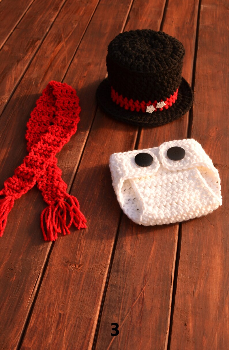 May include: A white crocheted nappy with two black buttons, a red crocheted scarf with tassels, and a black crocheted top hat with a red and white trim.