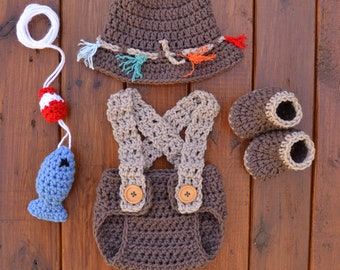 May include: A brown crochet baby outfit set, including a hat, overalls with wooden buttons, and booties. A blue crochet fish with a red and white bobber and white string are also included. The set is displayed on a wooden surface.