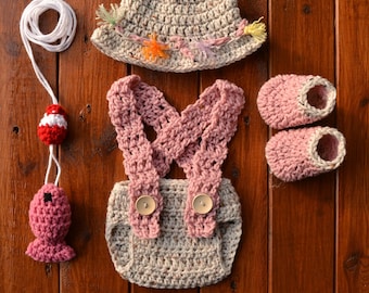 May include: A crocheted baby outfit set in beige and pink, including a hat, dungarees, and booties. The set features a whimsical fishing theme with a crocheted fish and fishing rod.