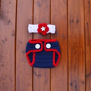 Puede incluir: Un conjunto de ganchillo que consta de una diadema blanca con una flor roja y un bot&oacute;n blanco en forma de estrella, y una funda para pa&ntilde;al azul marino con ribete rojo y dos botones blancos, con una textura de hilo tejido a mano.