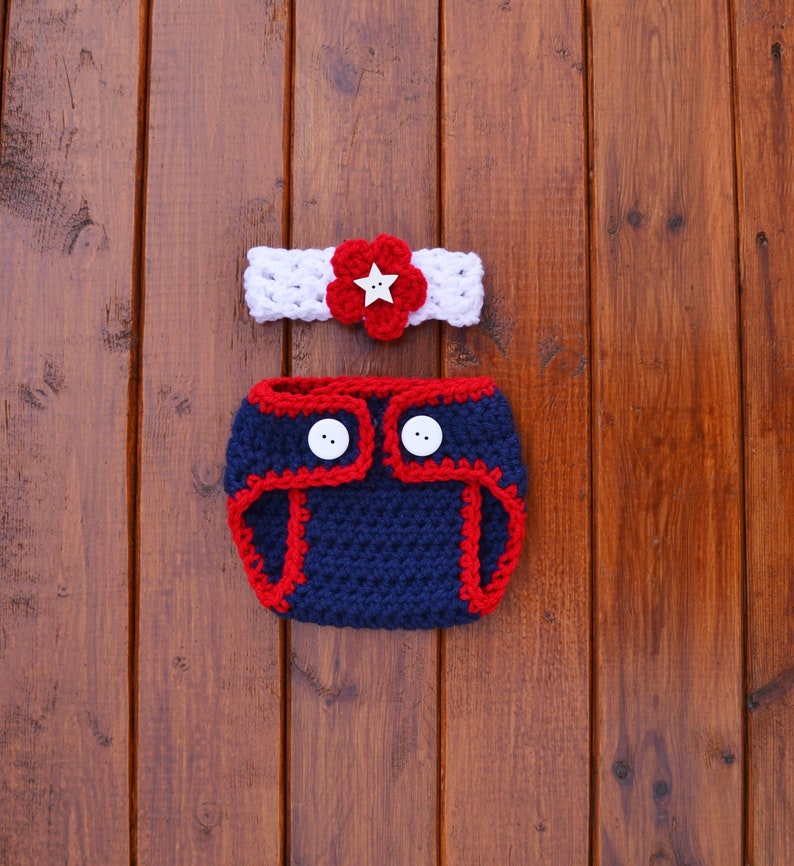 Puede incluir: Un conjunto de ganchillo que consta de una diadema blanca con una flor roja y un bot&oacute;n blanco en forma de estrella, y una funda para pa&ntilde;al azul marino con ribete rojo y dos botones blancos, con una textura de hilo tejido a mano.