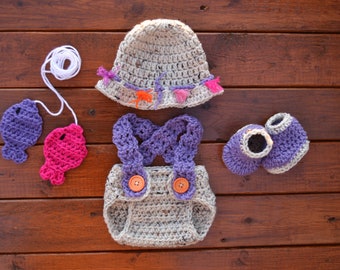 May include: A crocheted baby outfit set in grey, purple, and pink. The set includes a hat, a diaper cover, a pair of booties, and two fish-shaped toys.