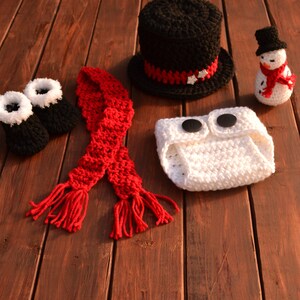 May include: A crocheted snowman outfit for a baby, including a black top hat, white nappy cover, red scarf, and booties.