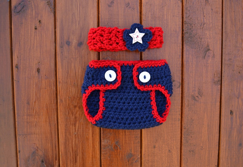 Puede incluir: Una funda de pa&ntilde;al de ganchillo azul marino con ribete rojo y dos botones blancos, acompa&ntilde;ada de una diadema de ganchillo a juego en azul marino y rojo con una flor azul oscuro y un bot&oacute;n blanco en forma de estrella con una peque&ntilde;a flor roja.