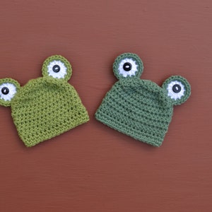 May include: Two green crocheted frog hats with white and black button eyes.