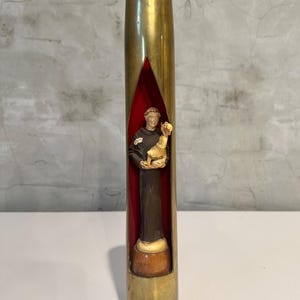 May include: A brass-coloured, cylindrical decorative piece with a red interior. Inside, a small statue of a figure holding a child is visible. The statue is set on a light-coloured base. The piece is displayed on a white surface.