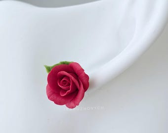 May include: A stud earring featuring a deep red rose with a small green leaf attached to the side. The rose has layered, curved petals that form a realistic flower shape. The text IRARUZHOVYCH is printed in small white letters below the rose.