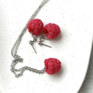 Könnte beinhalten: Ein Schmuckset bestehend aus einer silberfarbenen Kette und einem Paar Ohrsteckern, die jeweils mit einem realistischen, texturierten roten Himbeeranhänger versehen sind. Die Himbeeren sind mit kleinen silberfarbenen Ringen an den Metallteilen befestigt.