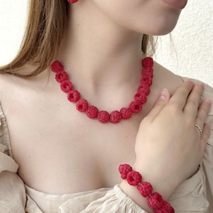 Op de afbeelding: Een sieradenset bestaande uit een ketting, een armband en een enkele oorbel, elk voorzien van meerdere kleine, getextureerde, felrode framboosvormige bedels. De ketting en armband zijn geregen met deze bessenkralen, terwijl de oorbel een enkele framboosbedel heeft die hangt aan een kleine zilverkleurige ring met heldere steentjes.