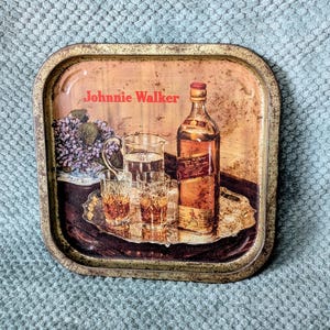 May include: Vintage metal serving tray with a Johnnie Walker advertisement. The tray features a bottle of whiskey, glasses, and a pitcher of water. The tray has a distressed, aged appearance.