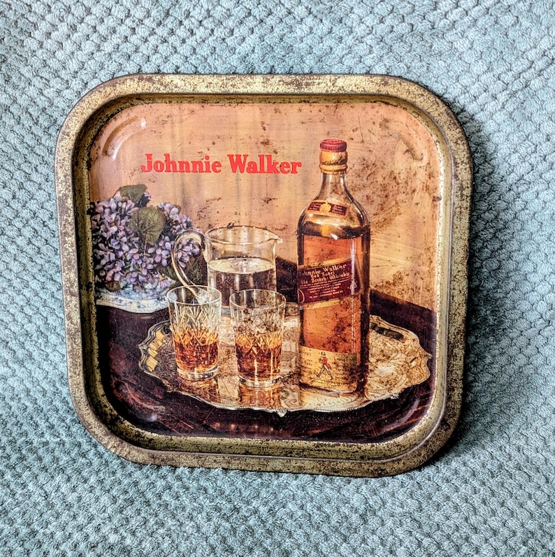 May include: Vintage metal serving tray with a Johnnie Walker advertisement. The tray features a bottle of whiskey, glasses, and a pitcher of water. The tray has a distressed, aged appearance.