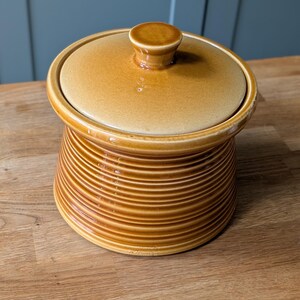 May include: A round, honey-colored ceramic storage jar with a matching lid. The body of the jar features a series of horizontal, raised ridges that wrap around the exterior, while the lid has a small, central knob handle for easy removal.