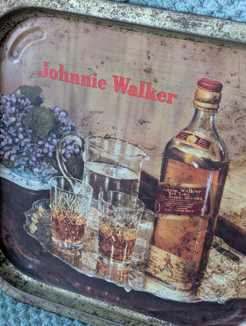 May include: Vintage metal tray with the text "Johnnie Walker" in red. The tray features a bottle of Johnnie Walker Red Label Scotch Whisky, two glasses of amber liquid, a pitcher of water, and a small bouquet of purple flowers.