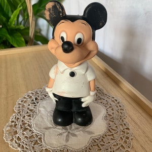 May include: Vintage Mickey Mouse figurine. The figure is wearing a white shirt with a black button, black shorts, and white gloves. The ears are black, and the face is tan. The figurine is standing on a gray doily.